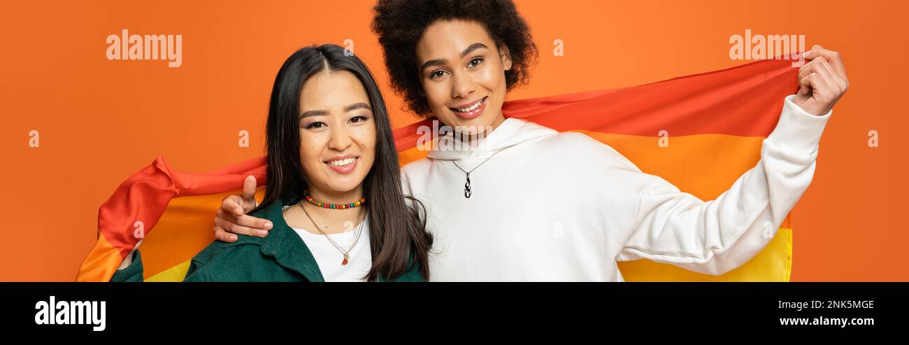 cheerful asian and african american women with lgbt flag looking at camera isolated on orange, banner,stock image Stock Photo