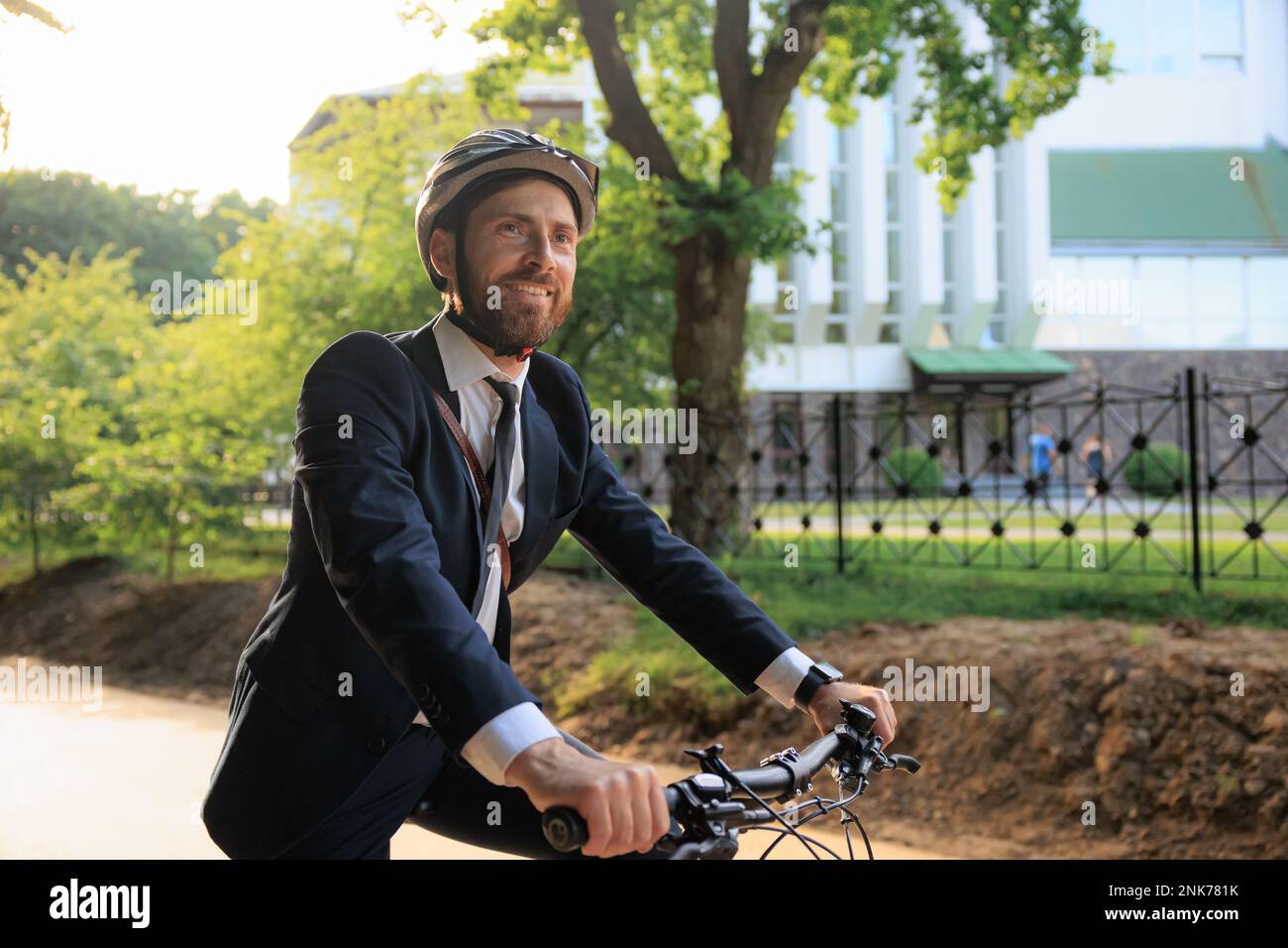 Happy male manager in black suit, riding on bicycle after workday in city. Side view of smiling worker in helmet looking ahead, enjoying trip, while d Stock Photo