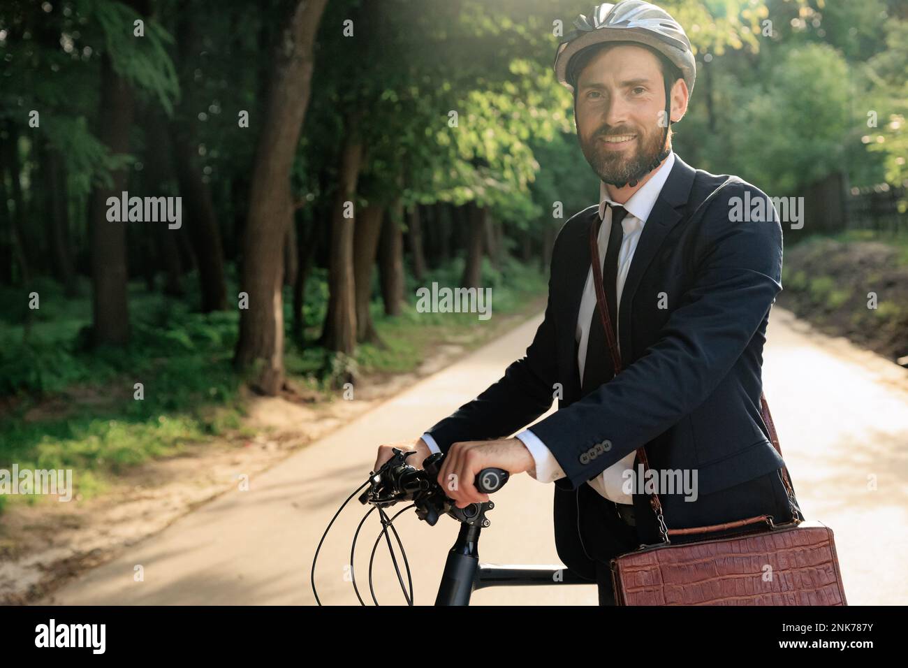 Confident businessman in helmet walking to work with bicycle in morning. Portrait of bearded entrepreneur in stylish suit standing with bike, while lo Stock Photo