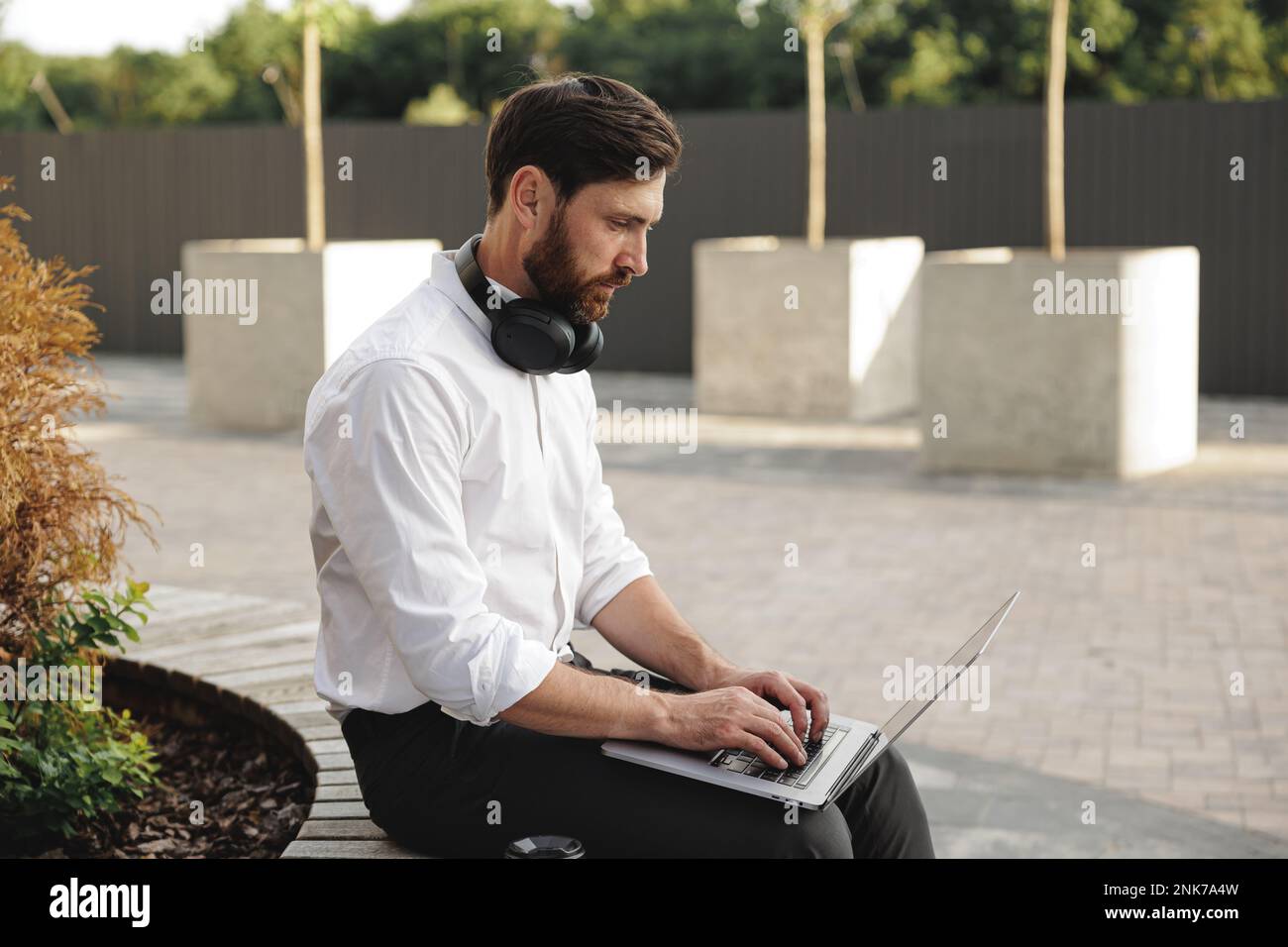 Concentrated bearded man typing while using laptop for redacting documents. Focused entrepreneur with headphones on neck working hard while sitting on Stock Photo