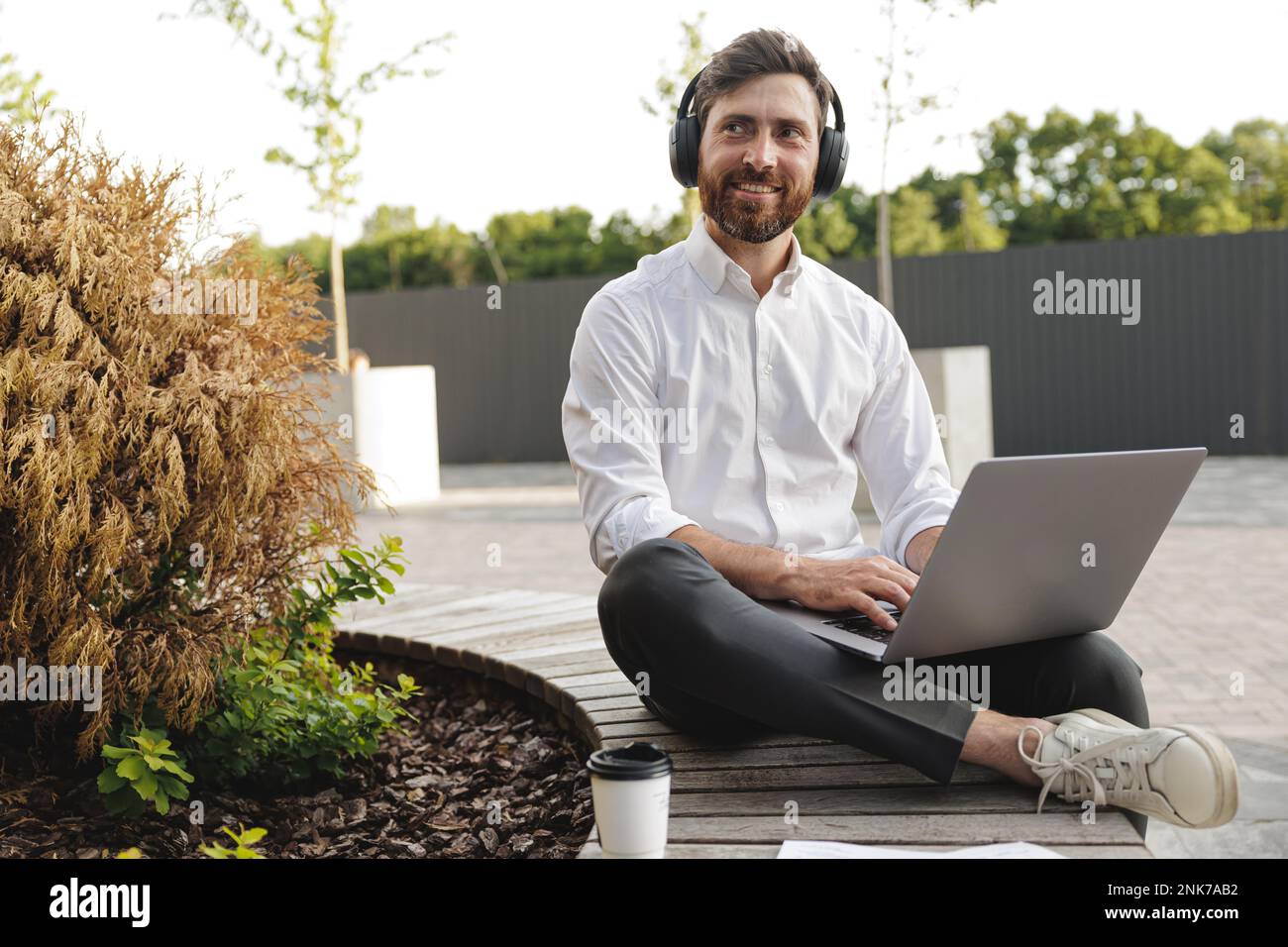 Joyful freelancer sitting on wooden bench with portable laptop on knees. Handsome man in black headphones looking aside with smile on face while worki Stock Photo