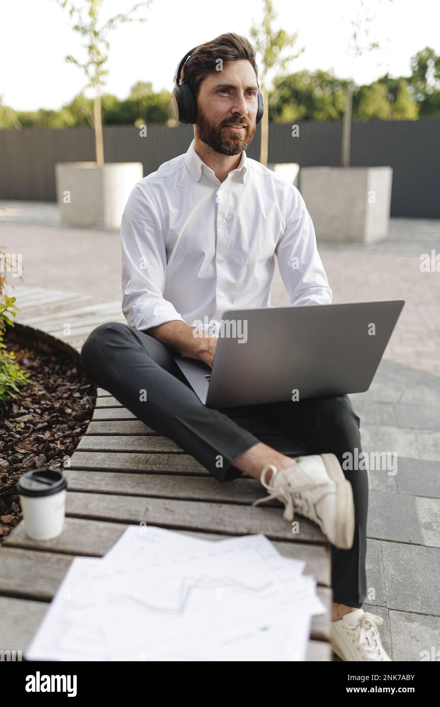 Stylish bearded man in wireless headphones looking aside and using portable laptop. Thoughtful freelancer sitting on bench near documents and coffee c Stock Photo