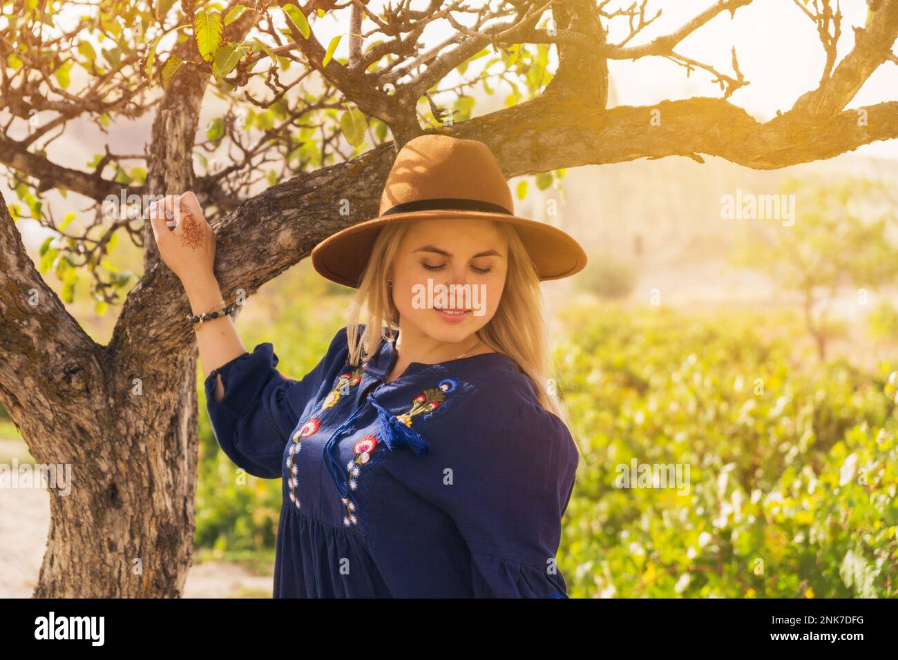 Beautiful blond hair woman wearing blue skirt and brown hat near tree in vineyard - plantation of grapevines. Spring dawn solar bright warm effect. Sl Stock Photo