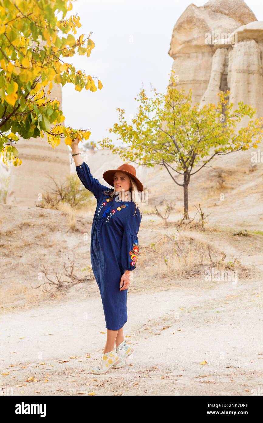 Traveling beautiful blond hair woman wearing authentic blue skirt and brown hat near autumn tree in Love Vally  in Goreme, Cappadocia, Turkey. Copy sp Stock Photo