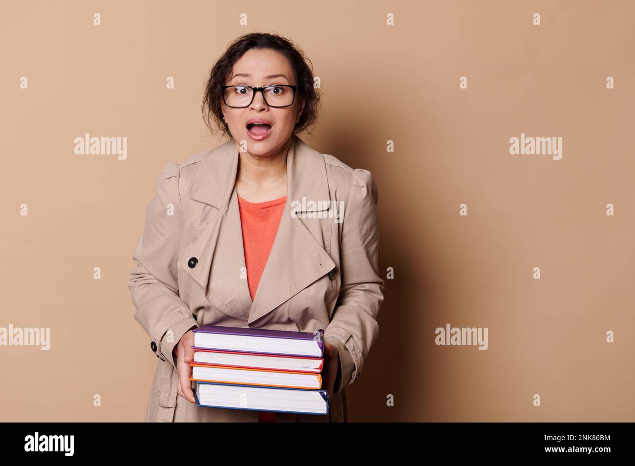 Astonished woman school teacher holds heavy volumes of hardcover books, expresses stupefaction at camera, cream backdrop Stock Photo
