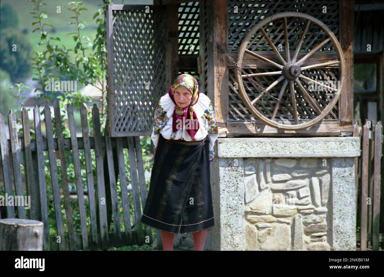 Straja, Suceava County, Romania, 1998. Elderly woman wearing traditional costume near a water well with wheel. Stock Photo