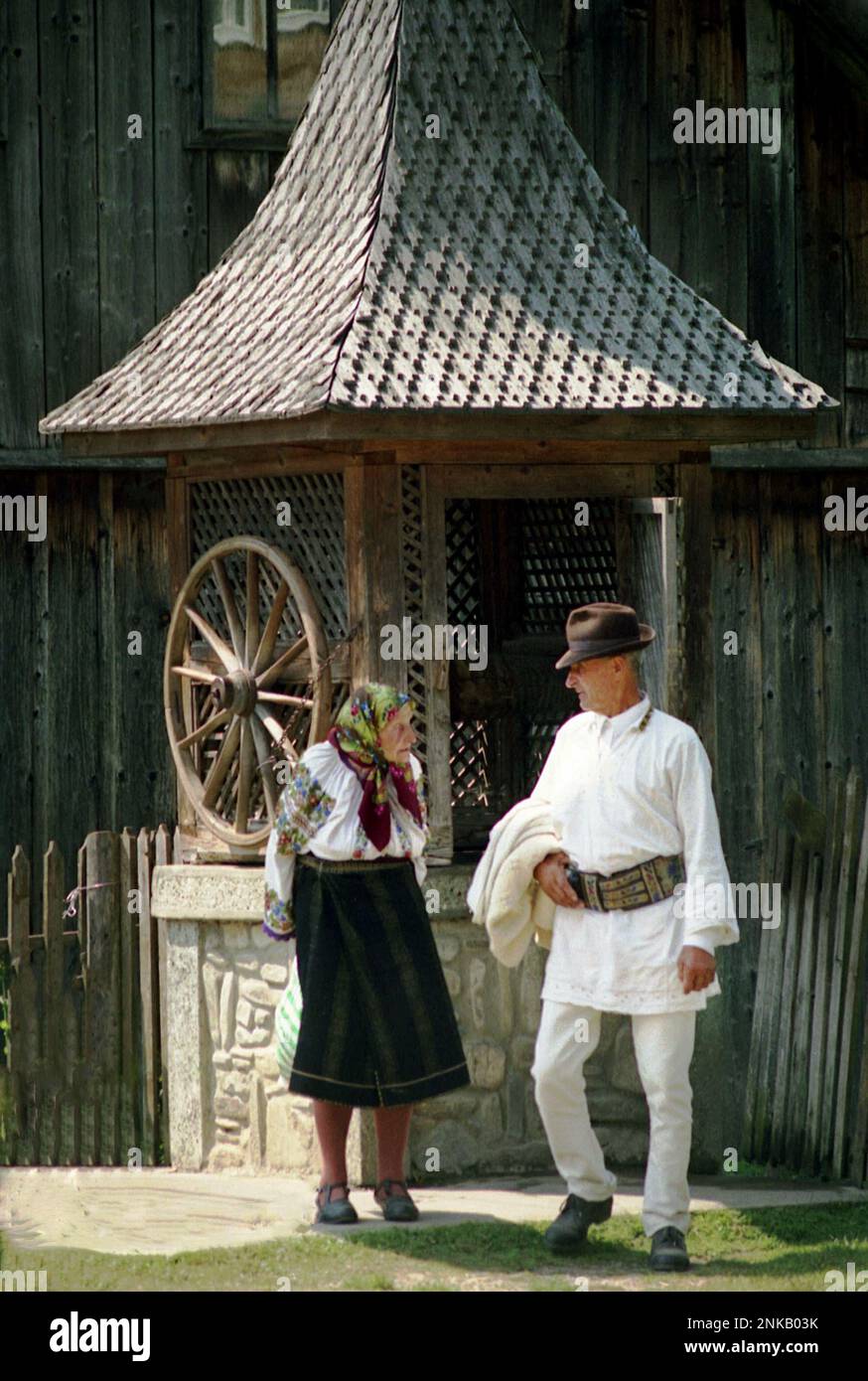 Straja, Suceava County, Romania, 1998. Elderly couple wearing traditional garments near a water well with wheel and wooden shingle roof. Stock Photo
