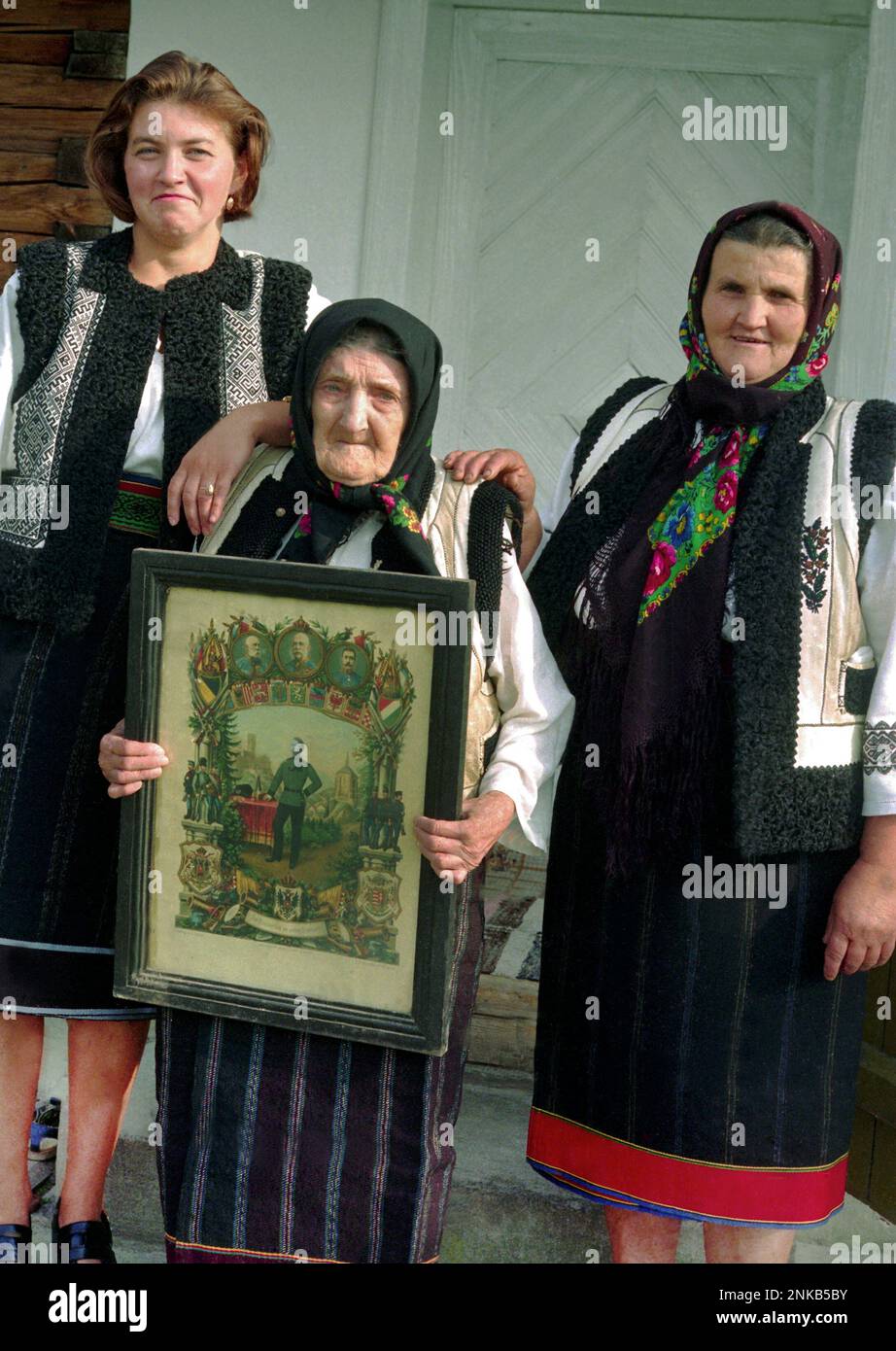 Suceava County, Romania, 1998. Three generations of women in a local family posing in their traditional costumes. The elderly woman is holding a framed printing portraying her husband as an infantry soldier in the Austro- Hungarian army. Stock Photo