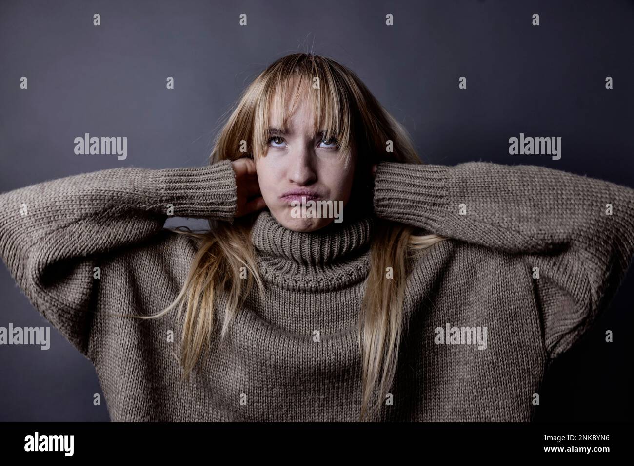 Young woman with long blond hair and a woollen jumper, arms crossed behind her head and face distorted, portrait, studio shot Stock Photo
