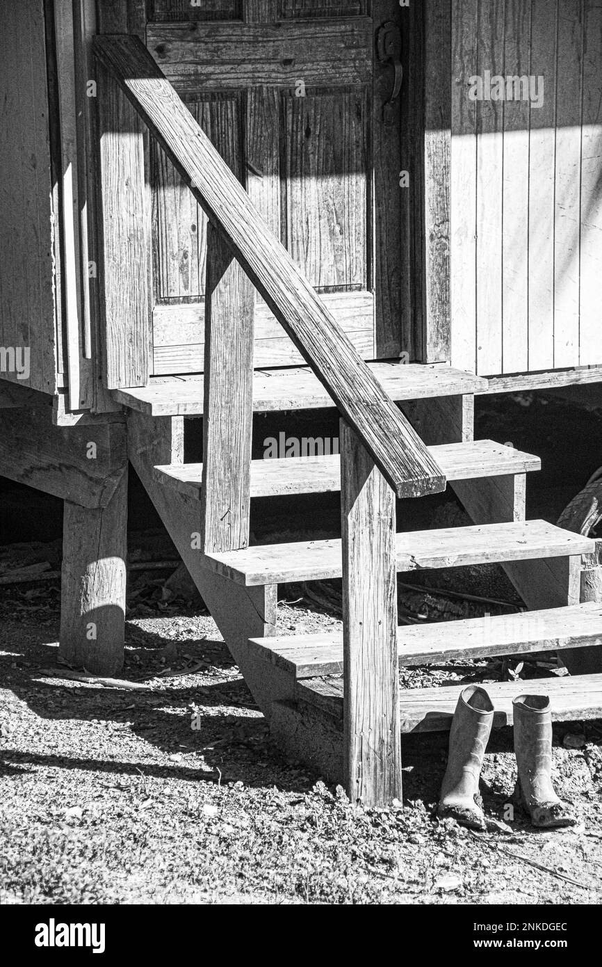 A black and white image of a pair of workboots by the side of stairs leading up to the door of a residence, Roatan, Honduras. Stock Photo