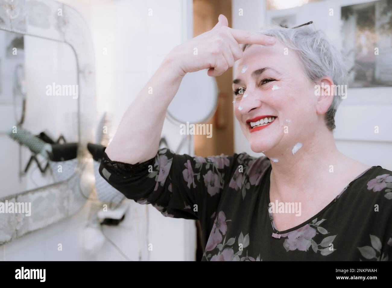 Happy senior woman applying moisturizer cream on face in bathroom Stock Photo