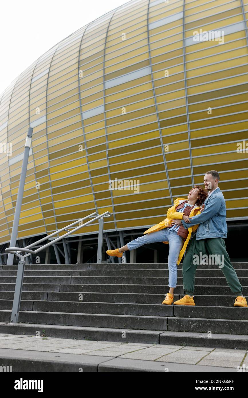 Playful woman falling over man on steps in front of stadium building Stock Photo