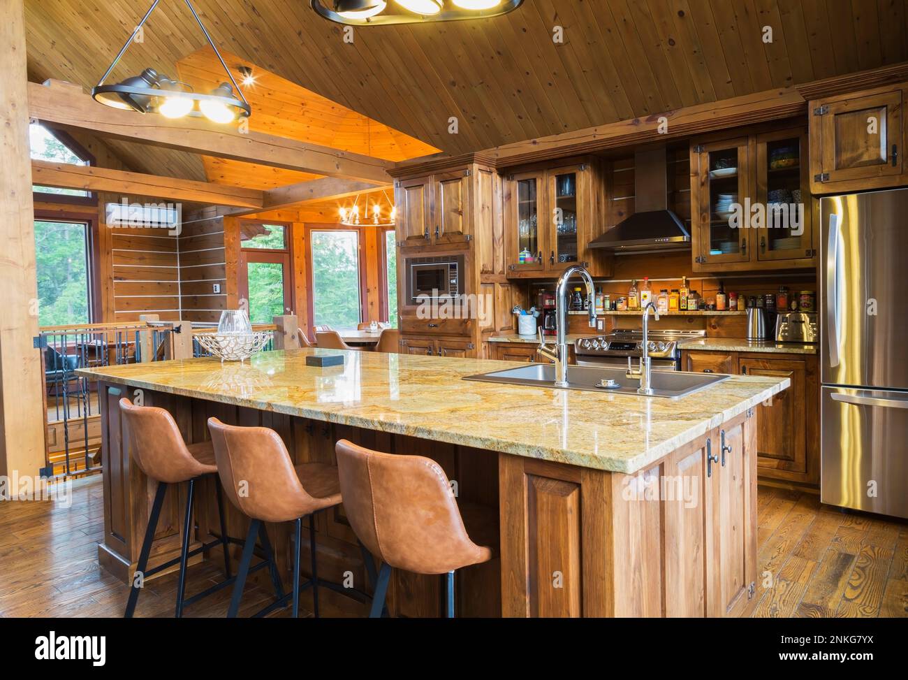 Pine wood cabinets with inlaid glass and island with yellow nuanced granite countertop and tan leather high chairs in kitchen inside milled log home. Stock Photo