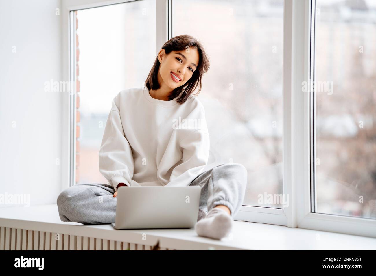 Happy young woman sitting with laptop on window sill Stock Photo - Alamy