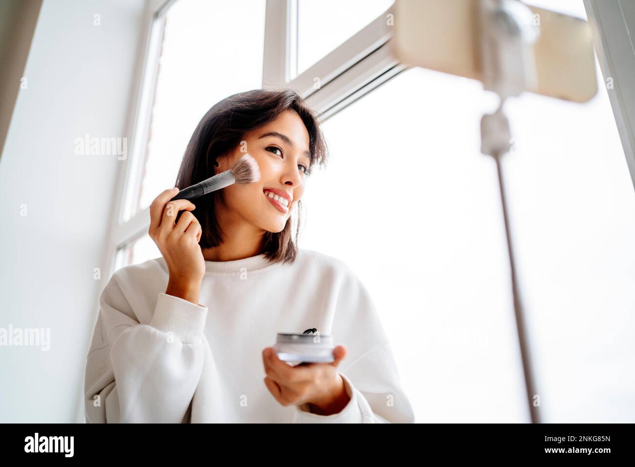 Young influencer applying blusher and filming through mobile phone at home Stock Photo