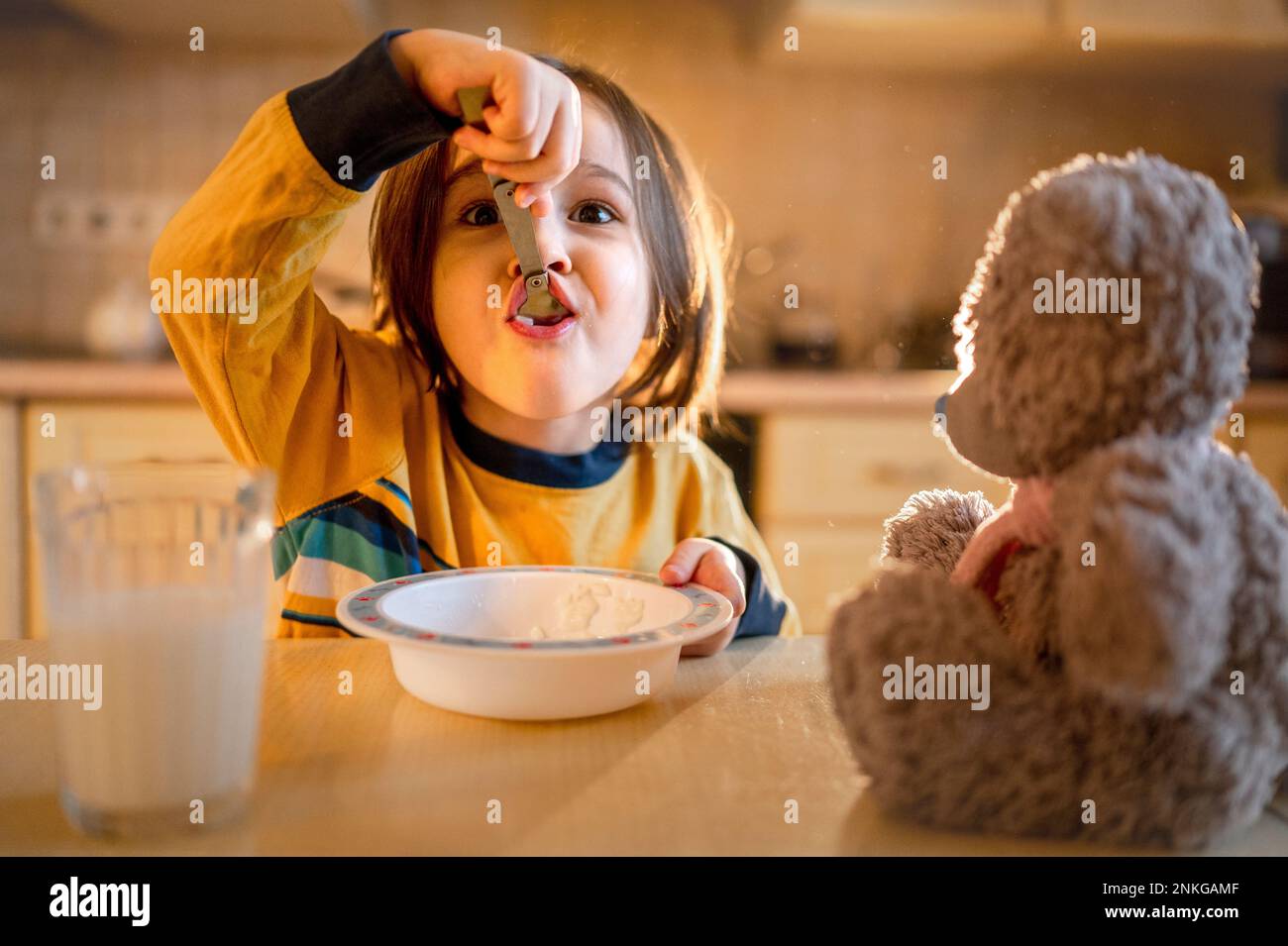 Boy eating porridge for breakfast by teddy bear on table at home Stock