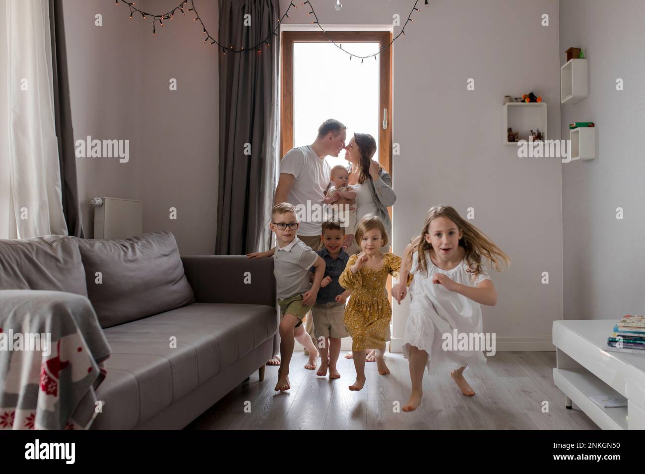 Children running in living room with parents rubbing nose in background Stock Photo