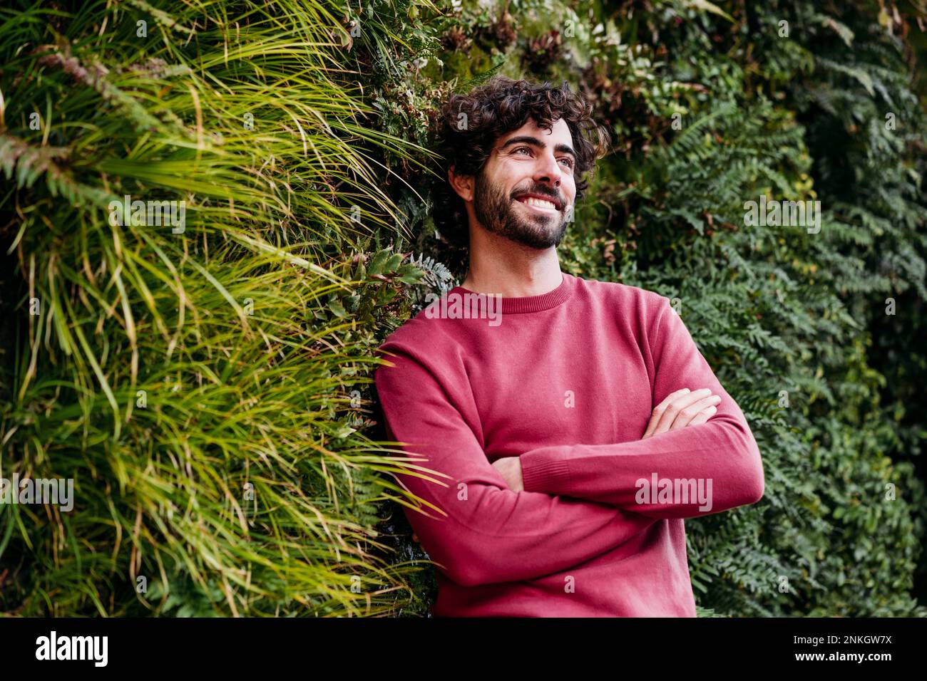 Thoughtful man with arms crossed leaning on plants Stock Photo