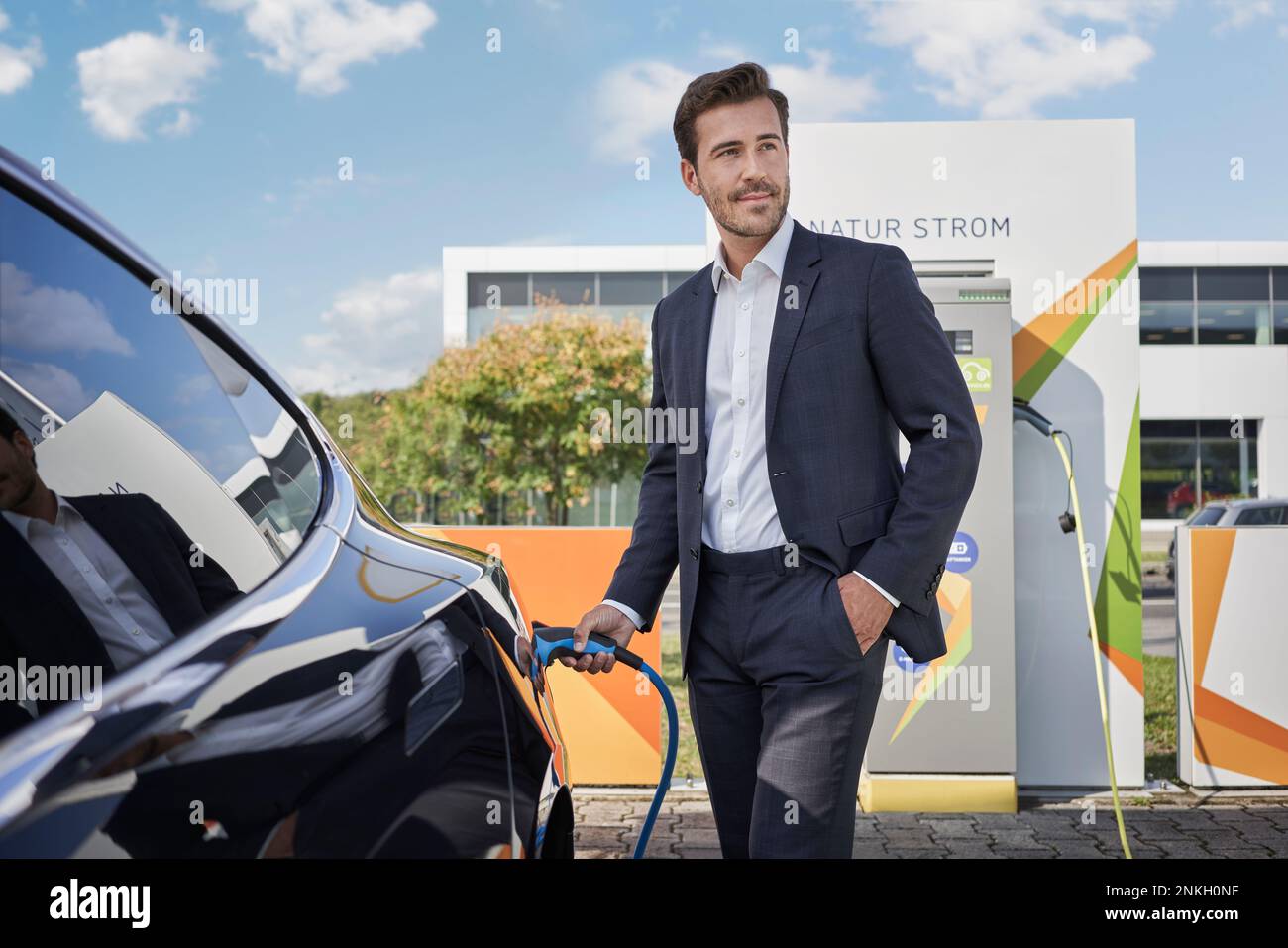 Smiling young businessman holding plug by car at electric vehicle charging station Stock Photo