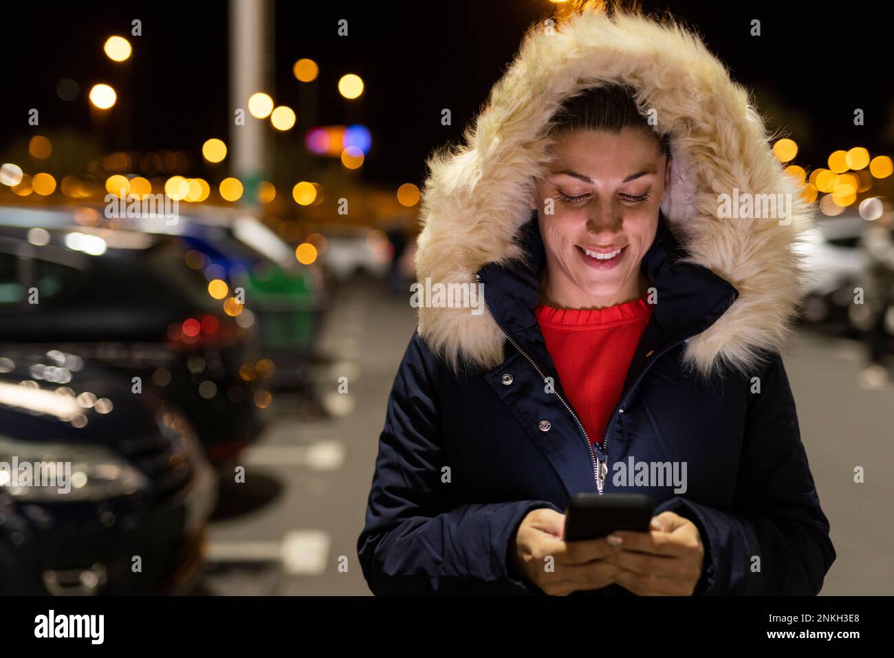 Happy woman wearing parka hood using smart phone at night Stock Photo