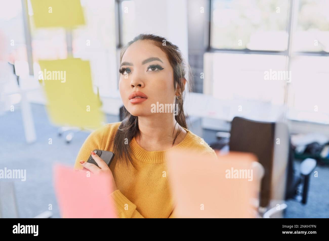 Thoughtful businesswoman with smart phone looking at adhesive notes on wall Stock Photo