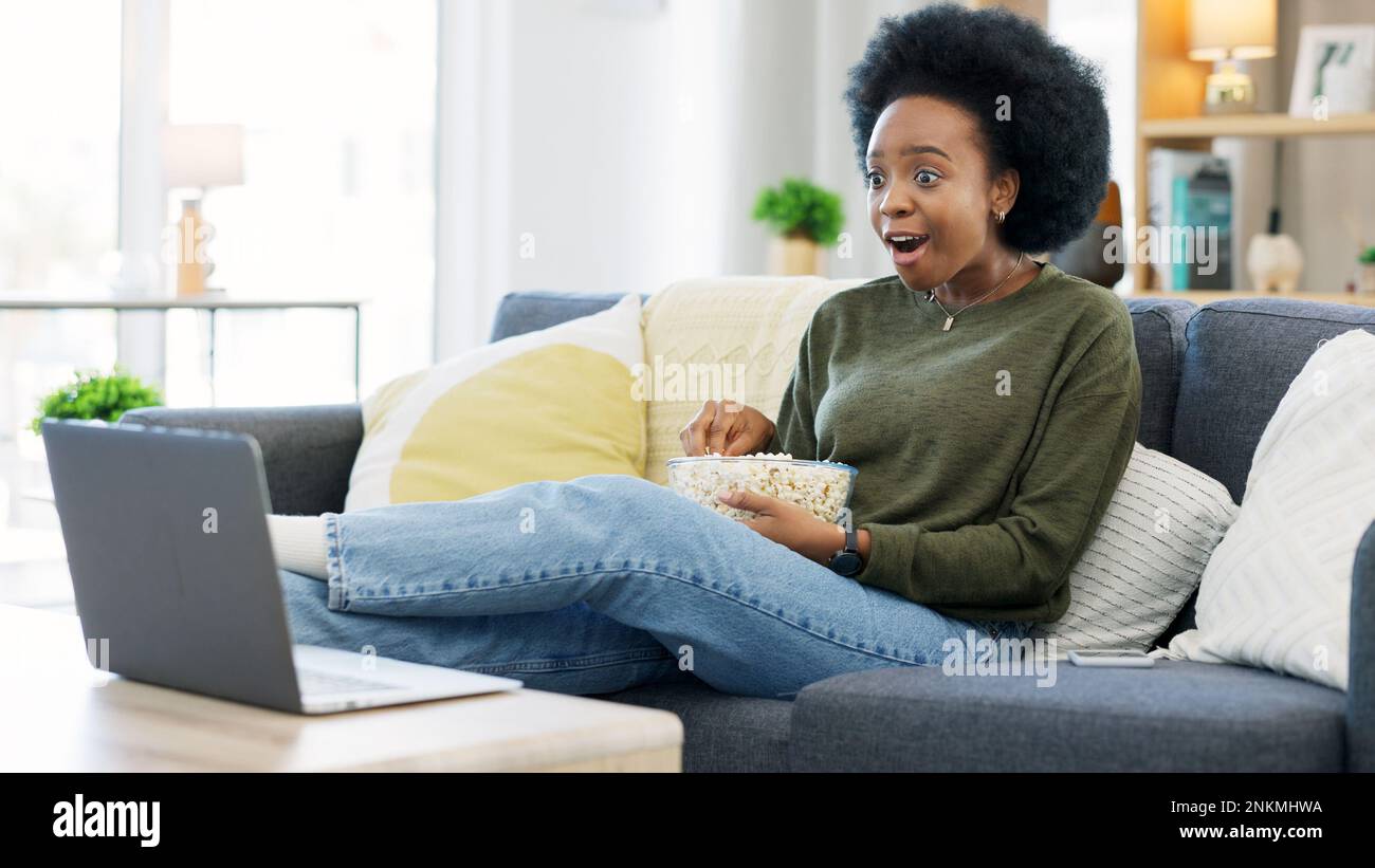 Happy african american woman streaming online movies on a laptop while snacking on popcorn and relaxing on a sofa at home. Black female enjoying a Stock Photo