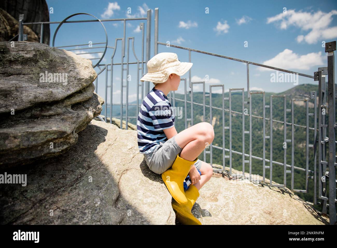 Young boy wearing hat and wellington boots sitting on rock looking at view on vacation Stock Photo