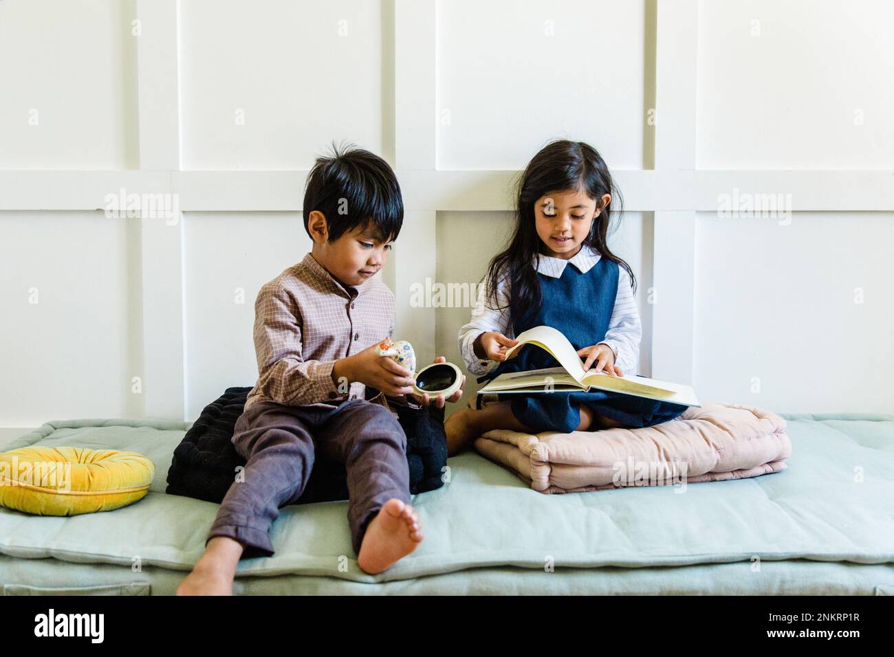 Two children sitting on cushions, girl reading book, boy holding object