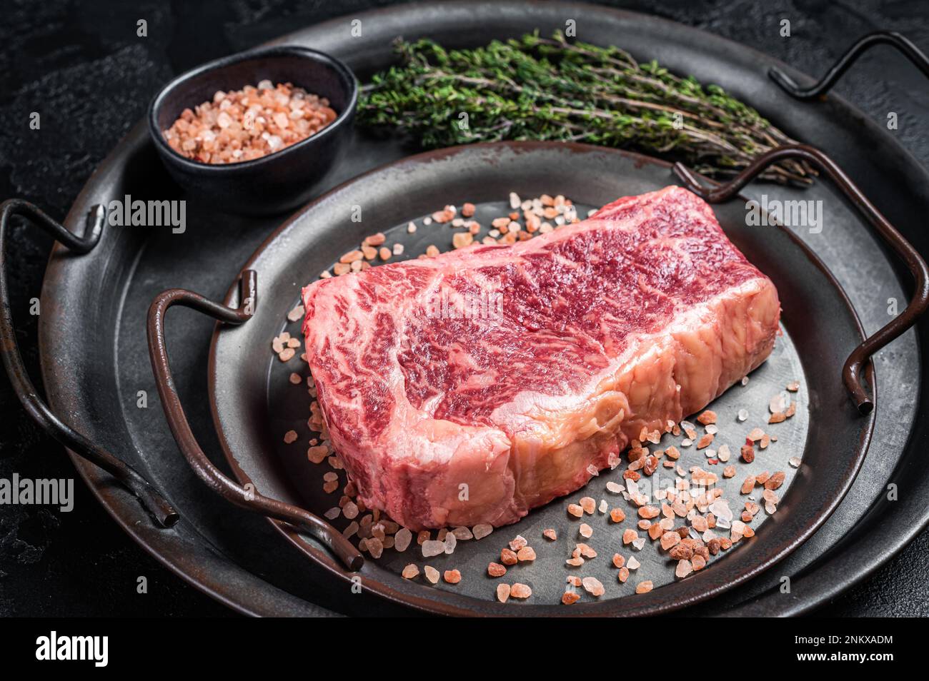 Fresh Raw Wagyu striploin or New york steak in a steel tray with herbs. Black background. Top view. Stock Photo