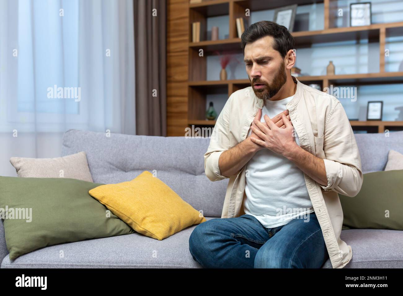 A young man is sitting on the sofa at home and holding his chest. Feels