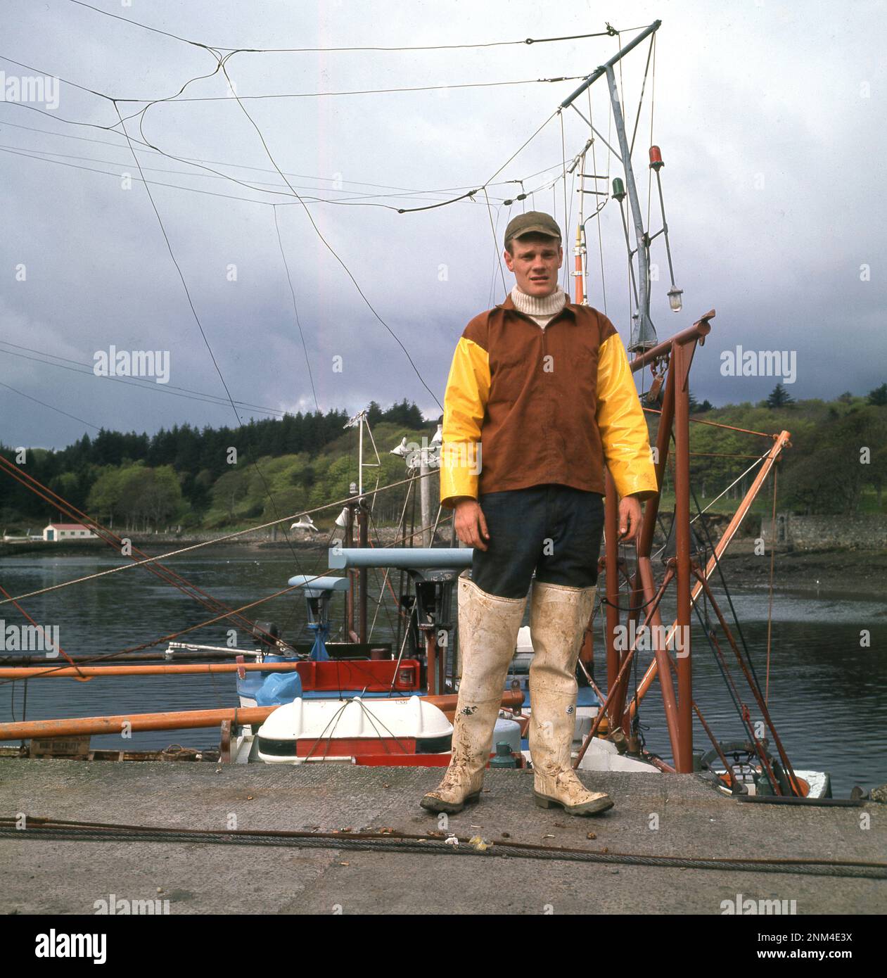 1960s, historical, a young adult fisherman in wadere, jacket and hat standing for a photo on the slipway at the harbour in Stornoway, on the Isle of Lewis in the Outer Hebrides, Scotland, UK Stock Photo