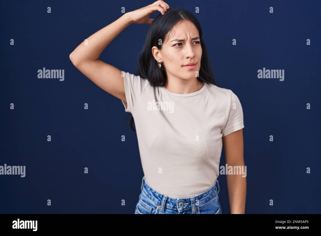 Young hispanic woman standing over blue background confuse and wondering about question. uncertain with doubt, thinking with hand on head. pensive con Stock Photo