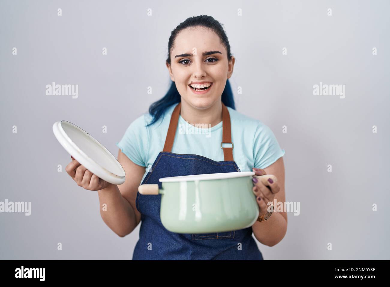 Young modern girl with blue hair wearing apron holding cooking pot