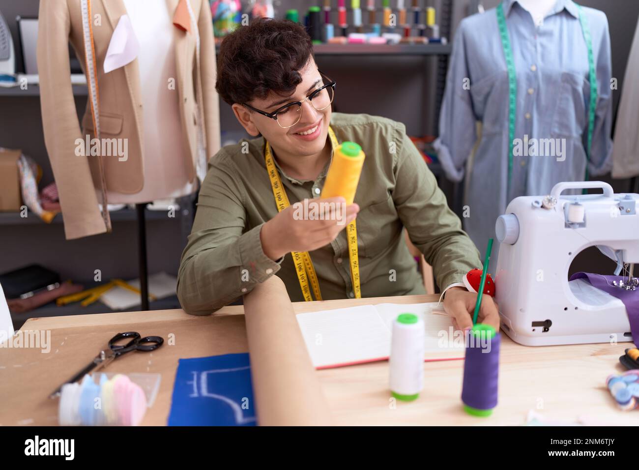 Non binary man tailor writing on notebook holding thread at atelier ...