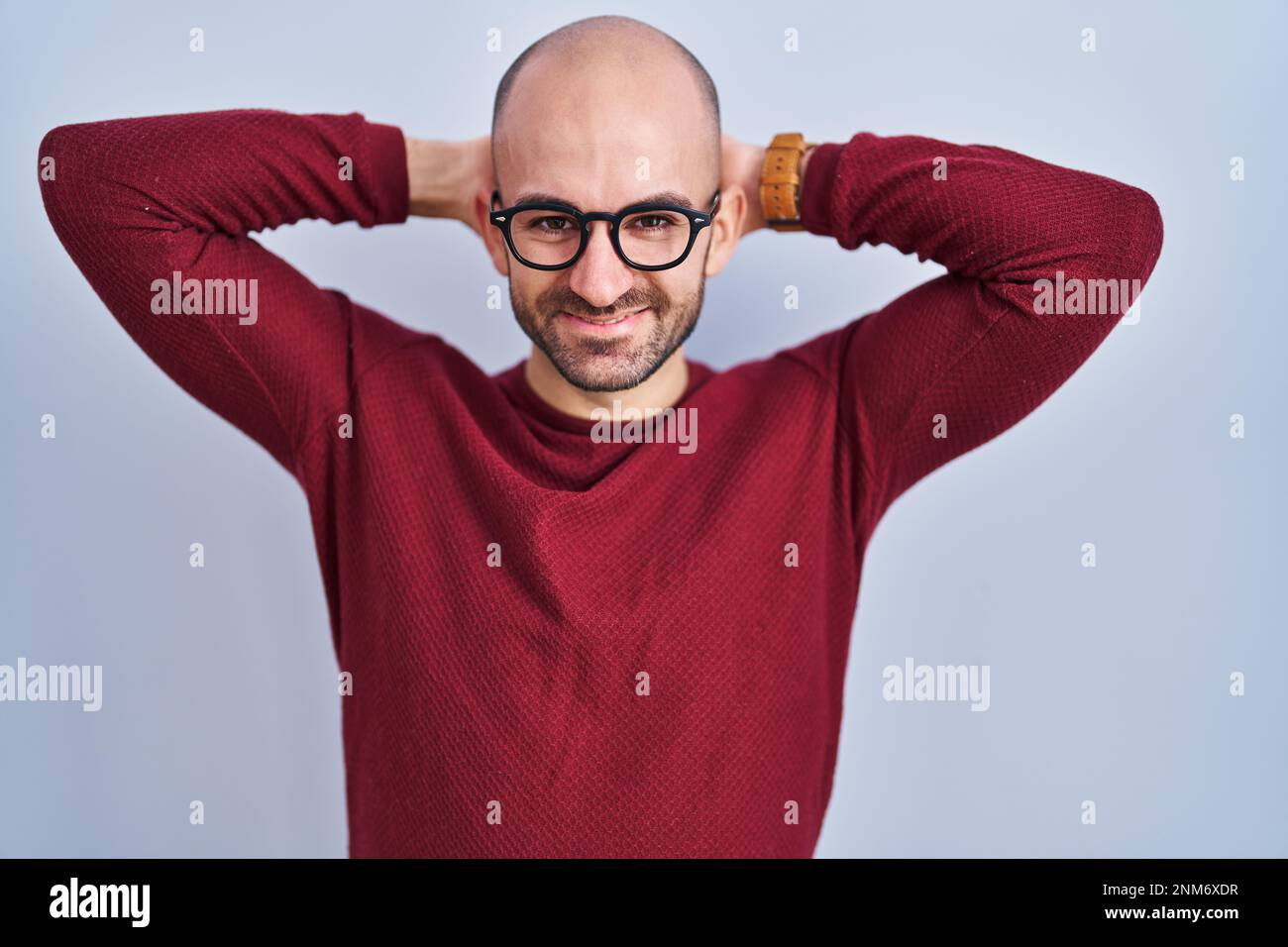 Young bald man with beard standing over white background wearing glasses relaxing and stretching, arms and hands behind head and neck smiling happy Stock Photo