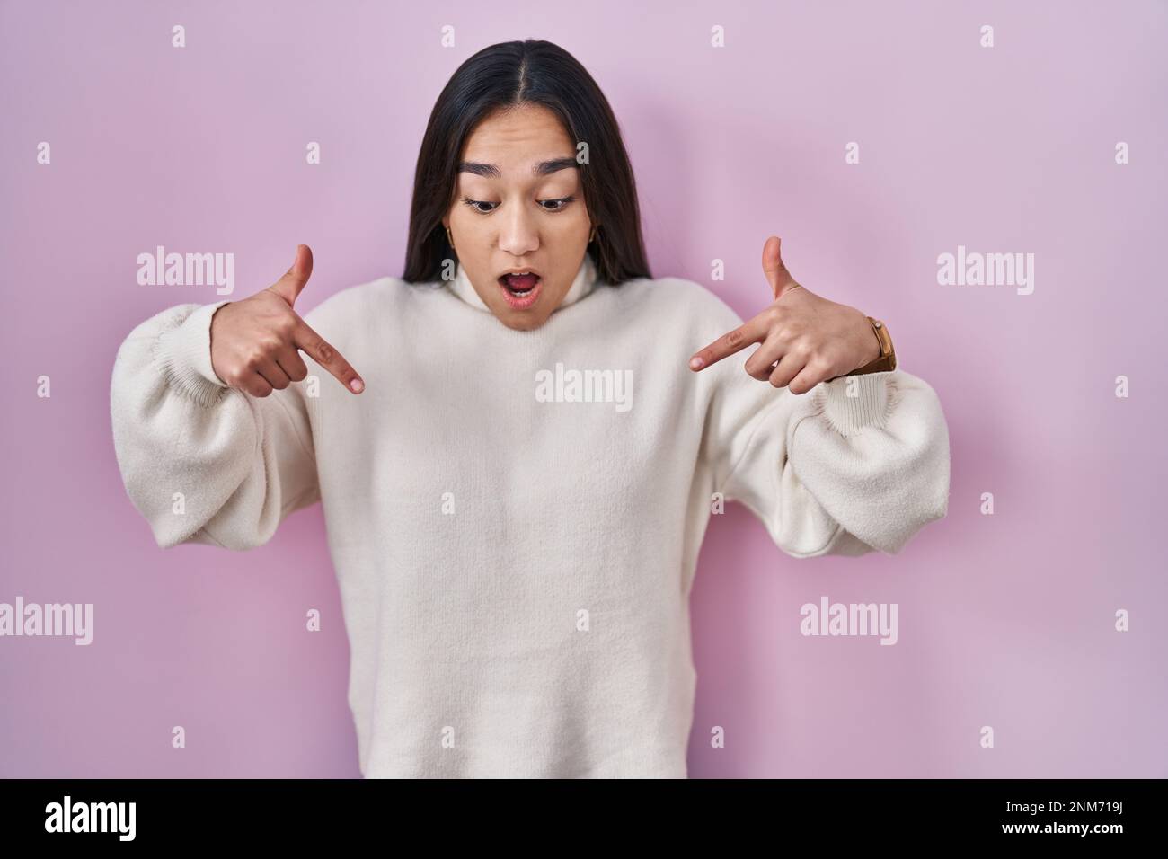 Young south asian woman standing over pink background pointing down with fingers showing advertisement, surprised face and open mouth Stock Photo