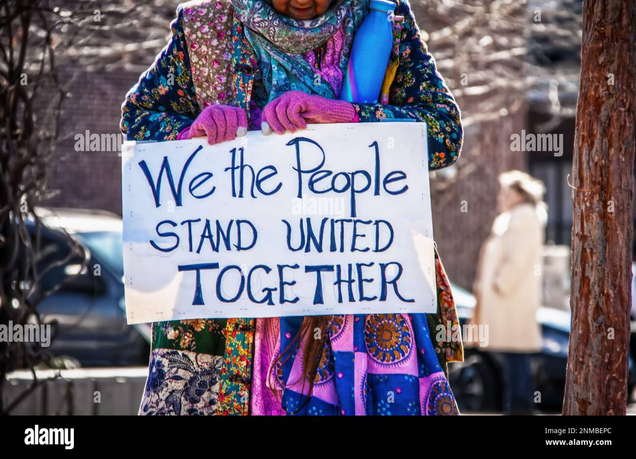 Older woman in colorful boho patchwork coat holds protest sign reading We the People stand United Together - blurred background Stock Photo