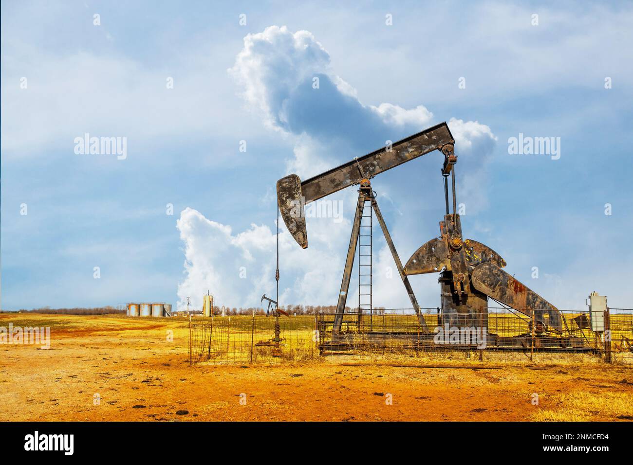Pump jack oil gas well on red soil with storage tanks on horizon under dramatic sky Stock Photo