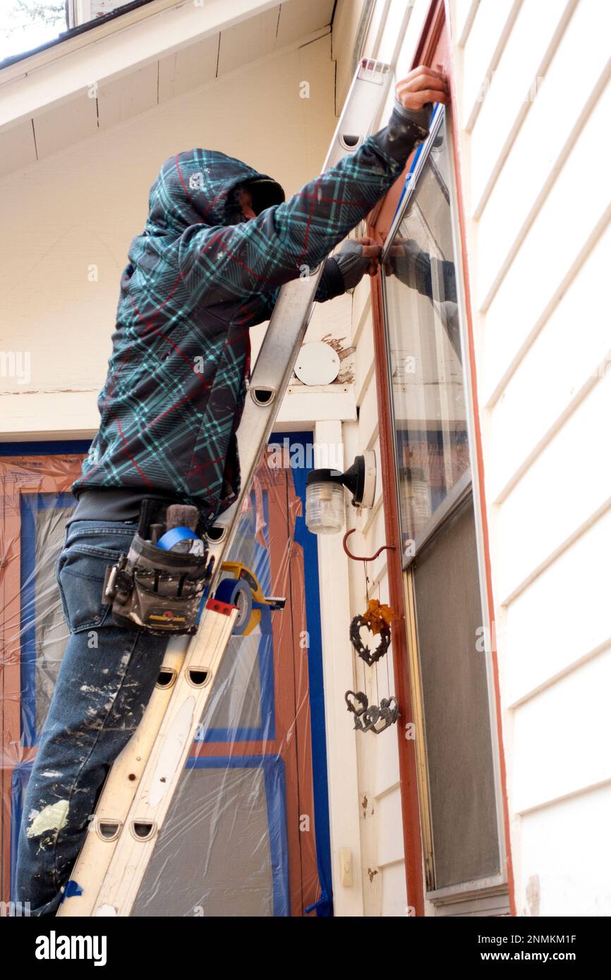 Painter using ladder reaching above window to apply blue edging tape. St Paul Minnesota MN USA Stock Photo