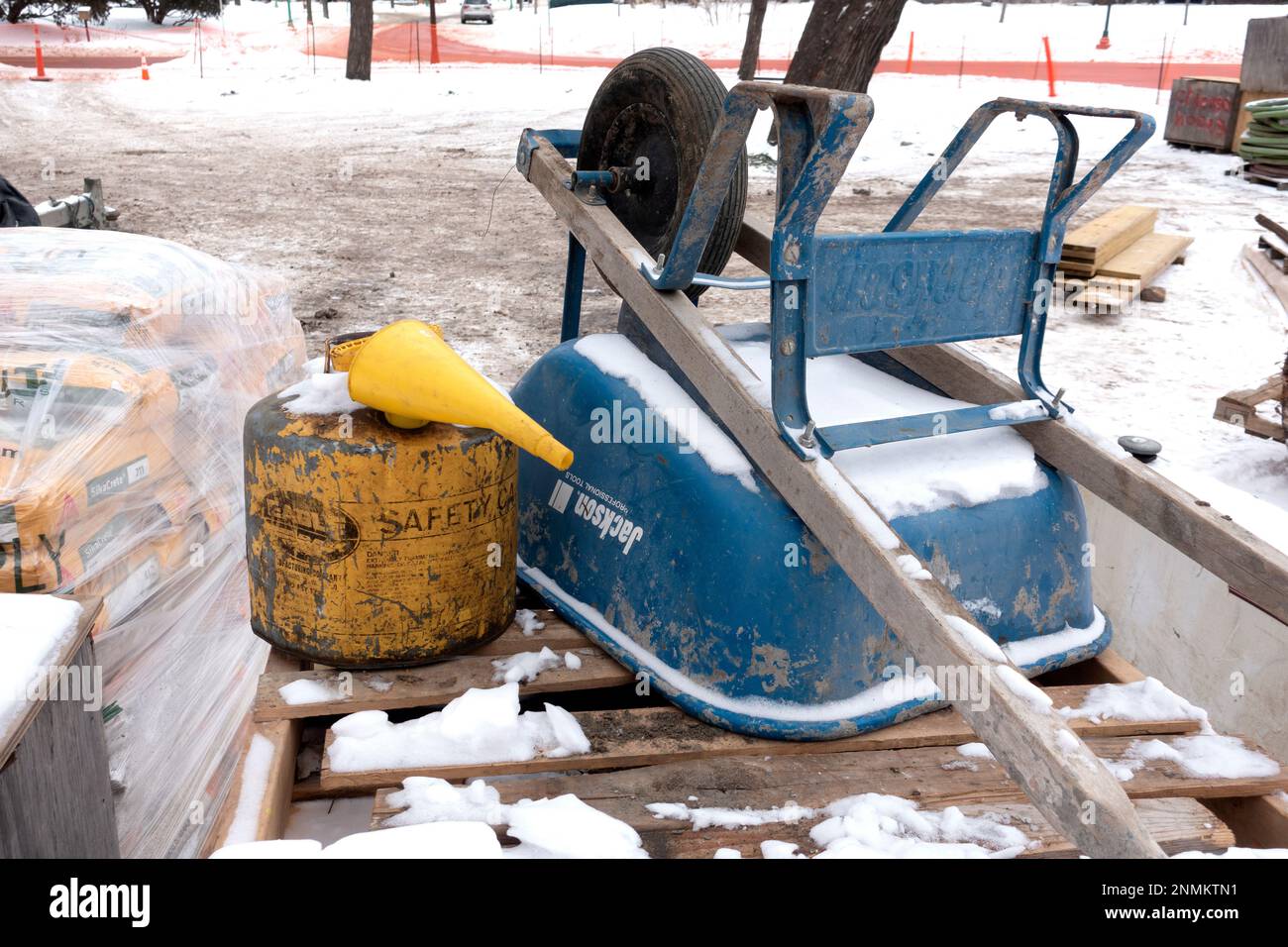 Yellow safety canister with funnel sitting on a wooden palate next to a blue wheelbarrow at a construction site. St Paul Minnesota MN USA Stock Photo