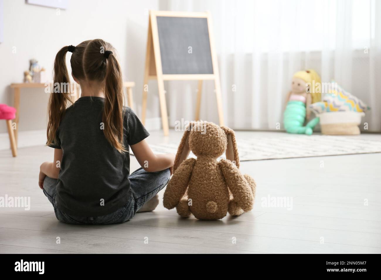 Lonely little girl with toy bunny sitting on floor at home, back view ...