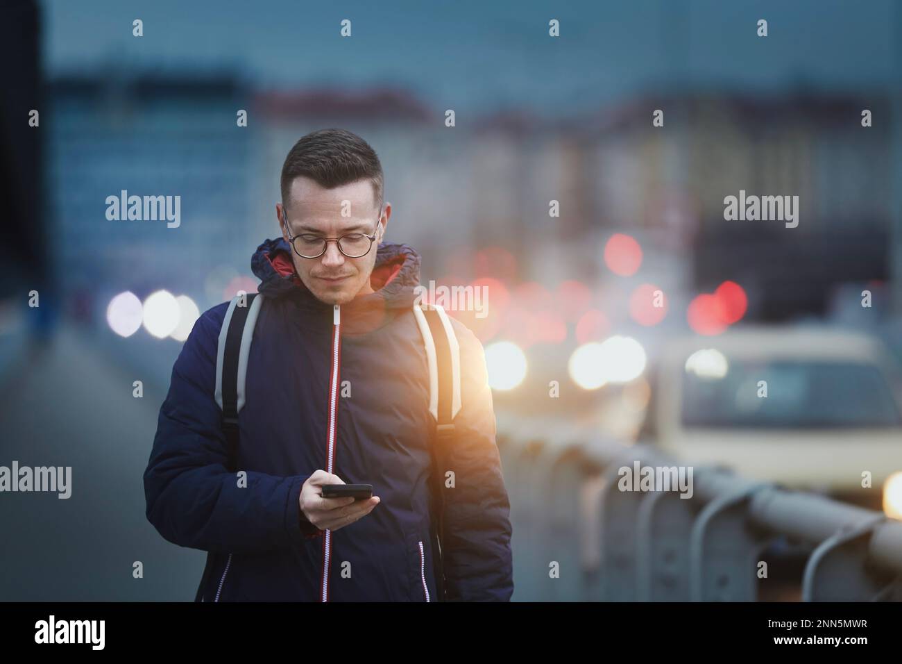 Man looking at phone while walking on busy city street. Urban scene with cars at twilight. Stock Photo