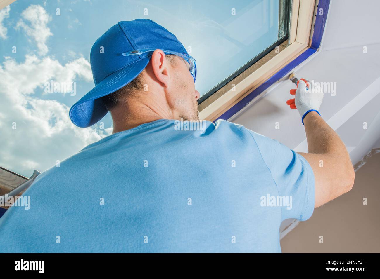 Closeup of Caucasian Contractor Carefully Painting Sloped Ceiling Around the Roof Window in White with a Brush. House Renovation Theme. Stock Photo