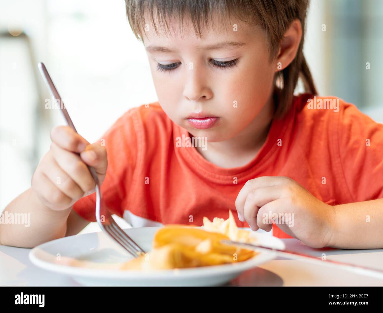 Little kid in red t-shirt eats pancakes with knife and fork. Curious boy with puzzled expression on face. Tasty pastry for breakfast. Stock Photo