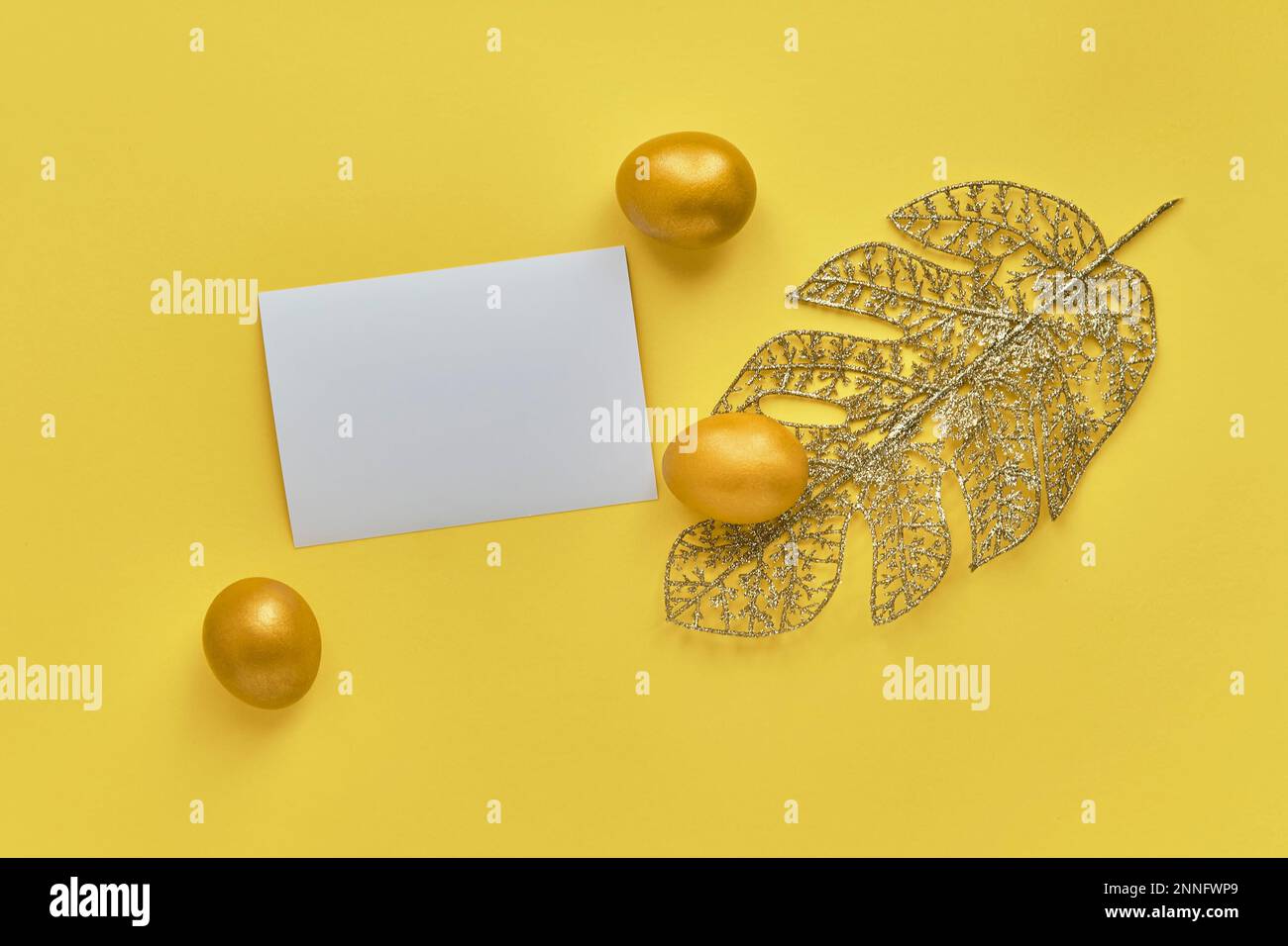 Gold eggs, monstera leaf from wire, blank paper, yellow background. Stock Photo