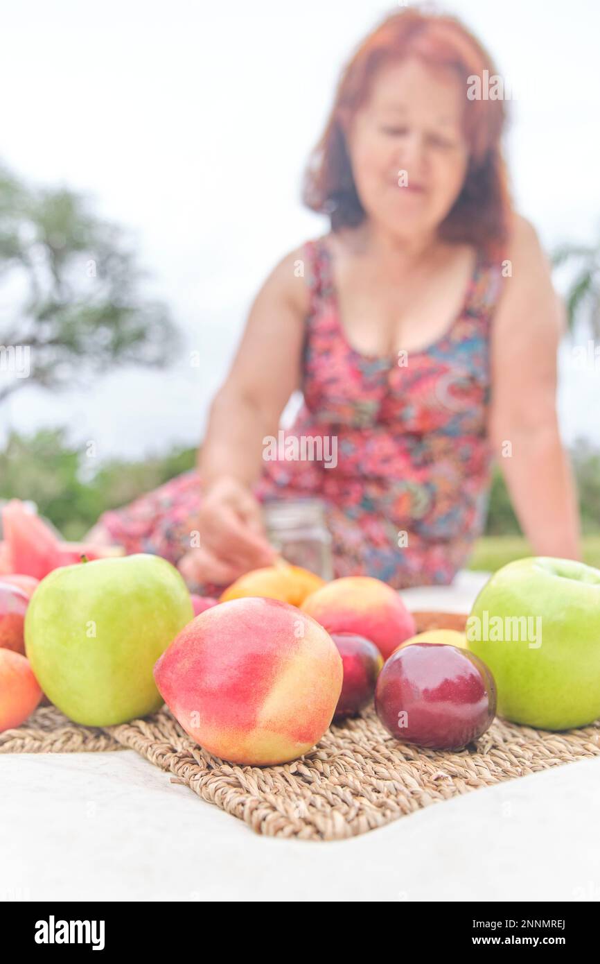 Mature woman having a picnic breakfast with a variety of fresh fruits, apples, peaches, tangerines, plums, watermelon. Focus on them. Concepts: health Stock Photo