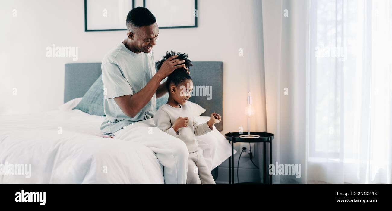 Black dad styling his daughter’s Afro hair into a bun while sitting on a bed. Father gives his daughter a new hairstyle. Dad and daughter spending the Stock Photo