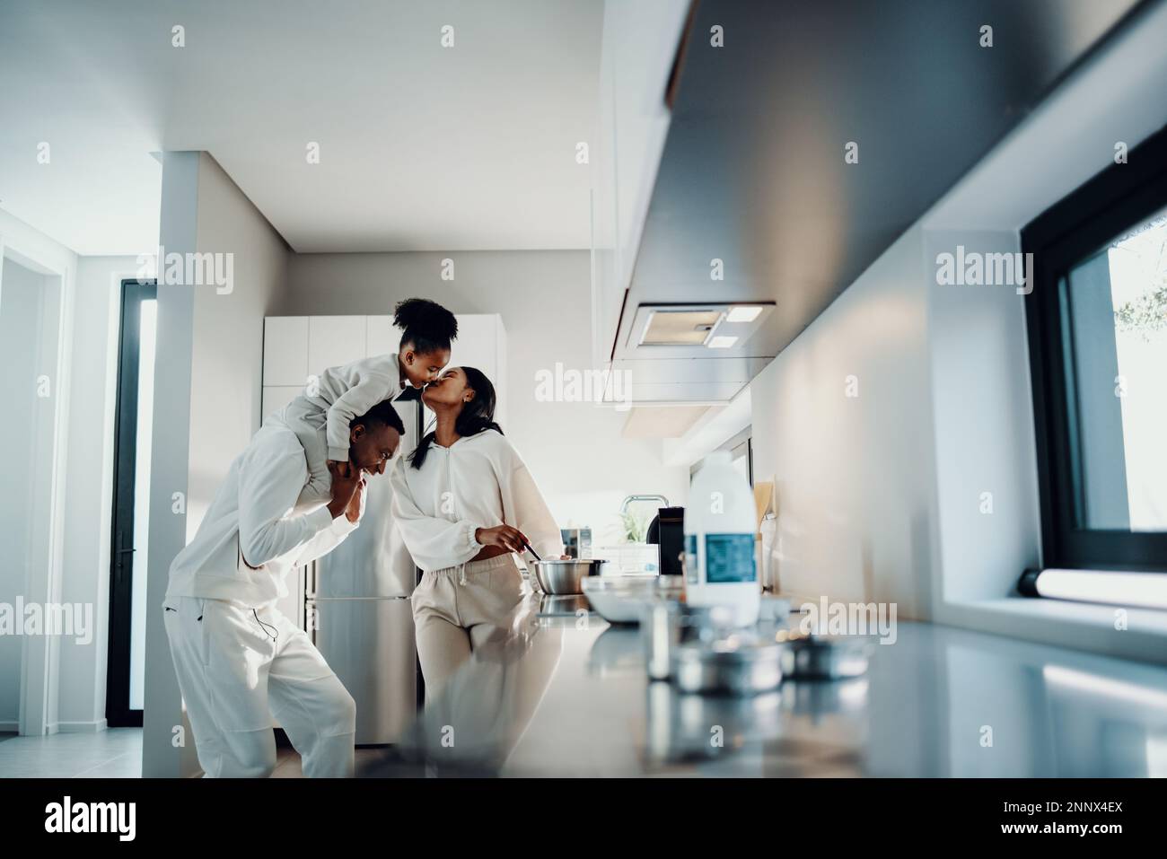 Family fun at breakfast time. Mom kisses her daughter from dad’s shoulders. Family having playful moments as mom prepares breakfast in the morning. Stock Photo