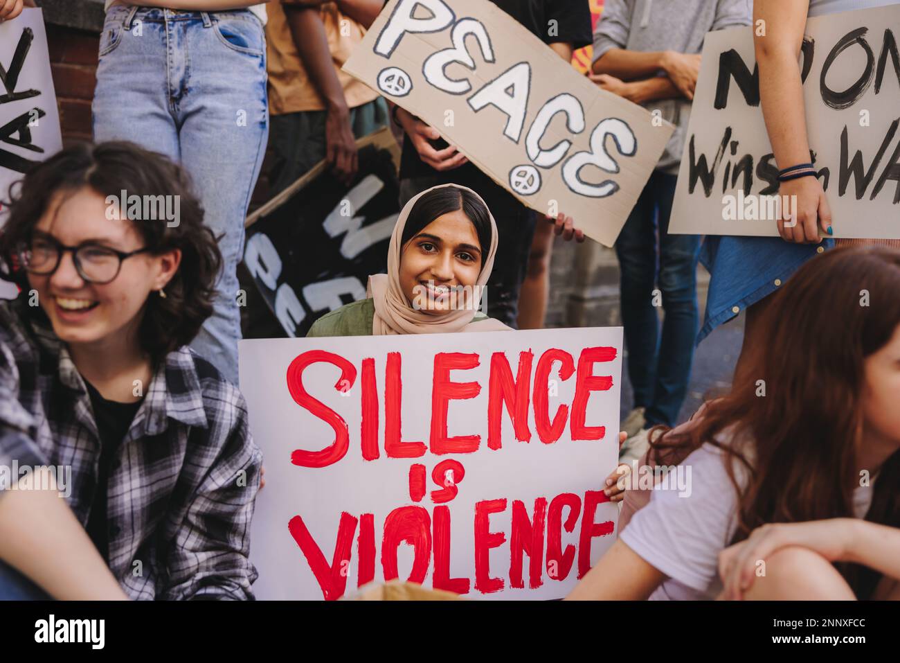 Happy Muslim girl smiling at the camera while holding an anti-violence poster. Diverse teenagers protesting against war and ethnic violence. Youth act Stock Photo