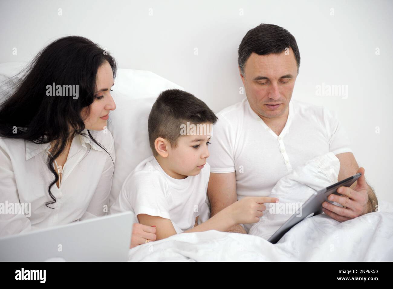 banner latest technology family husband wife and child sitting watching movie on laptop on bed relaxing together white clothes chatting online with relatives mom dad and son laughing smiling rejoicing Stock Photo